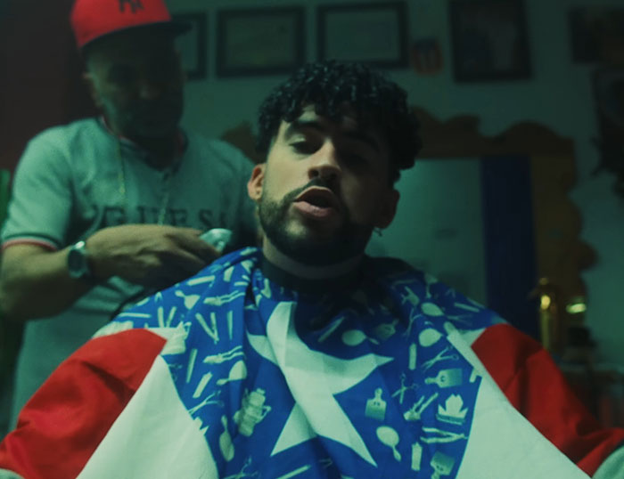 Bad Bunny getting a haircut wearing a Puerto Rican flag cape with a barber in the background in a dimly lit room.