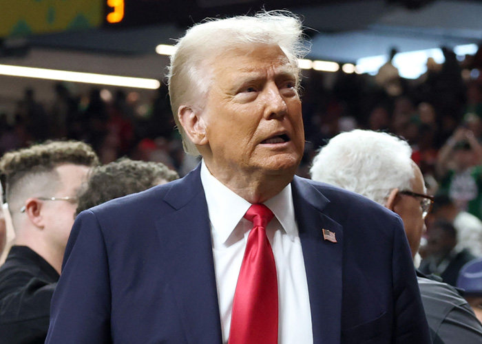 Donald Trump in a navy suit and red tie at a crowded event, reacting amid discussions of Bad Bunny&rsquo;s Super Bowl halftime show.