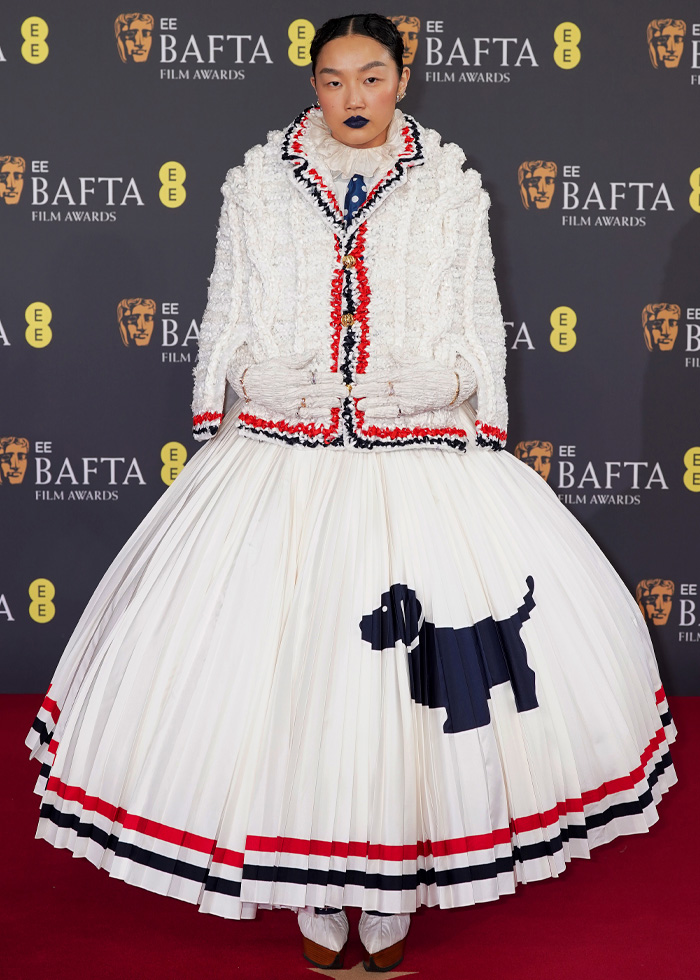 Model wearing a white textured jacket and pleated skirt with dog motif, standing on the BAFTA 2026 red carpet in standout outfits.