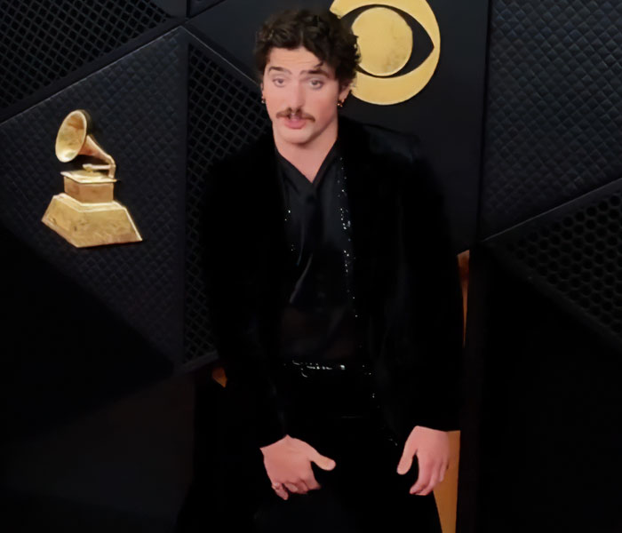 Male celebrity in black outfit posing awkwardly at the 2026 Grammy Awards with gold gramophone backdrop visible.