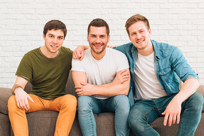 Three young men sitting closely on a couch, smiling, representing family dynamics with son's mistress and clueless wife.