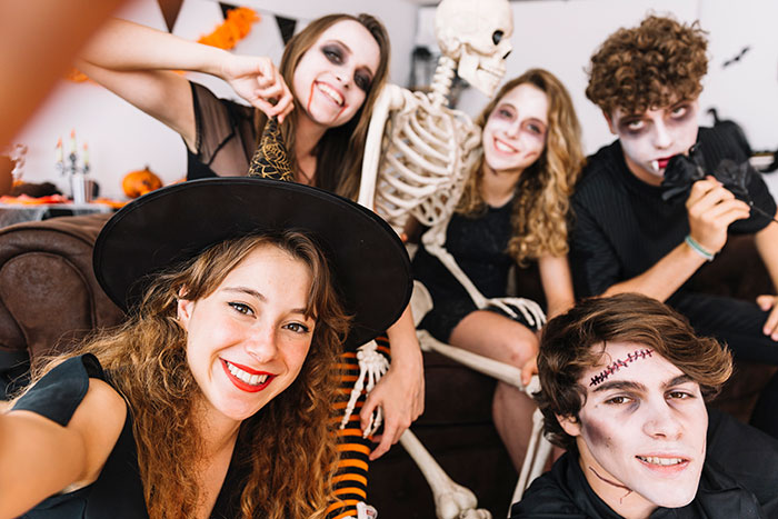 Group of friends in Halloween costumes with younger sister trouble theme, smiling and posing for a selfie at a party.