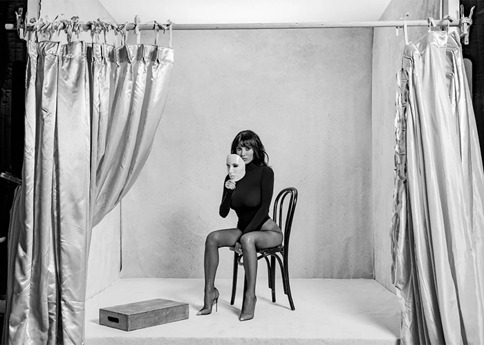 Bianca Censori sits on a chair in a black outfit holding a white mask in a minimalist black and white photo studio setting.