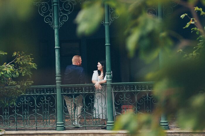 Couple having a first date conversation on an ornate balcony surrounded by greenery, capturing awkward encounters.