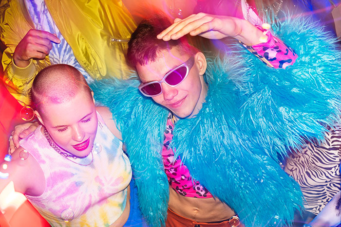 Two young people wearing colorful outfits and dancing at a lively party with bright neon lights and energetic vibes.