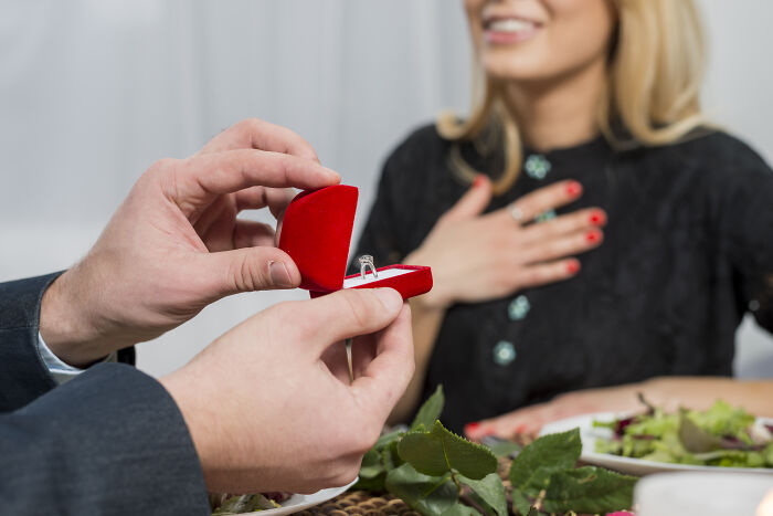 Man holding an open red ring box proposing to a smiling woman during a romantic dinner scene with engagement focus.