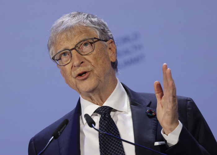 Bill Gates speaking at a conference, addressing audience with a raised hand and wearing glasses and a dark suit.