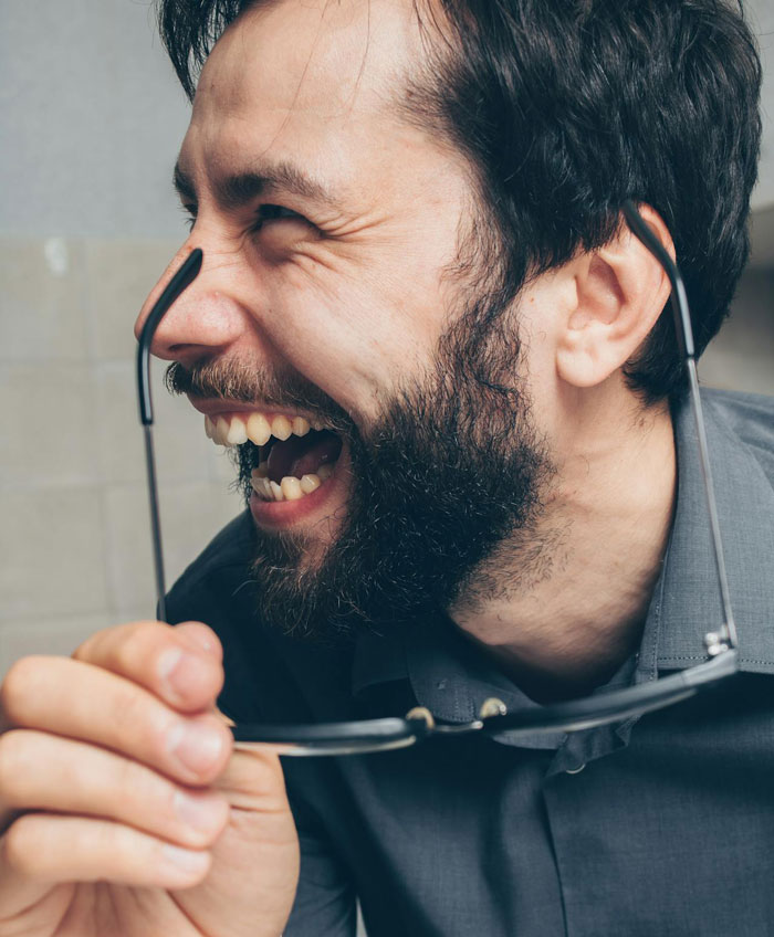Man laughing while holding glasses, reacting to boss commenting on his low weight in a casual setting.