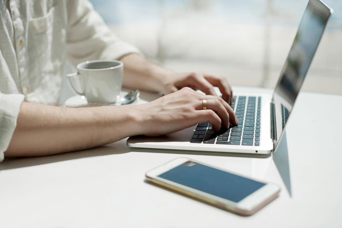 Person typing on laptop keyboard with coffee cup and smartphone nearby, illustrating report review by boss.