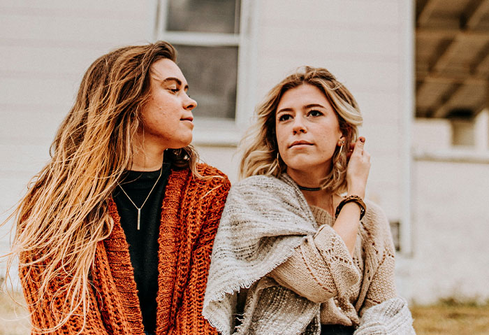 Two women sitting outside, one looking unsure while the other appears to confess, illustrating cheating and denial conflict.