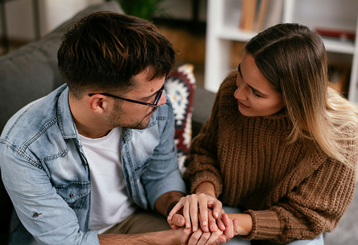 Young woman and man holding hands on a couch, showing tension and emotional conflict after cheating confession and denial.