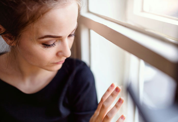 Woman looking out window, appearing confused and troubled after best friend&rsquo;s cheating confession and boyfriend&rsquo;s denial.