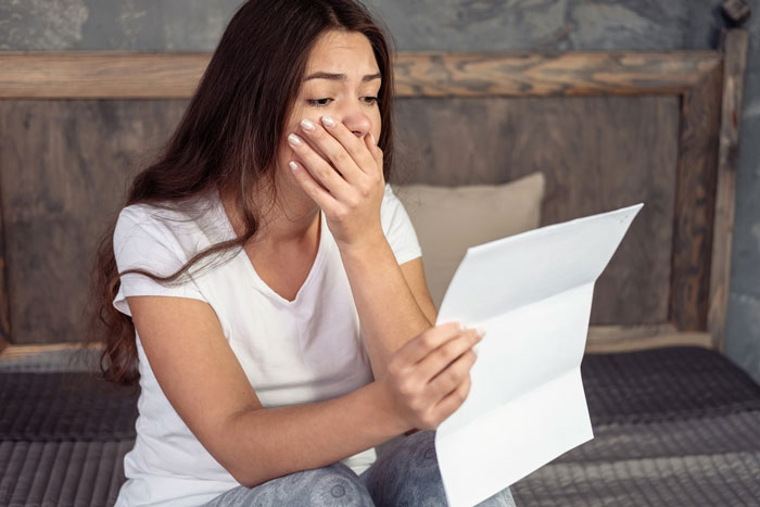 Young woman sitting on bed, covering her mouth in shock while reading a letter about a cheater breaking her heart.
