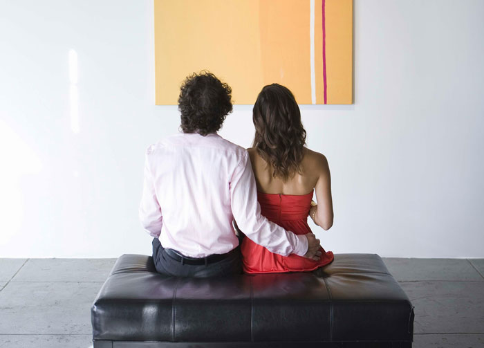 Couple sitting on bench in a museum setting with boyfriend holding girlfriend&rsquo;s waist, hinting at fake proposal love moment.