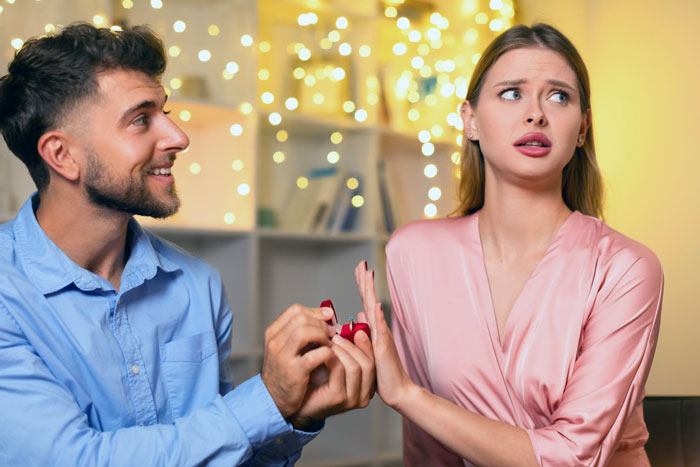 Young man making a fake proposal with a ring while the woman shows discomfort at a love museum setting with warm lights.