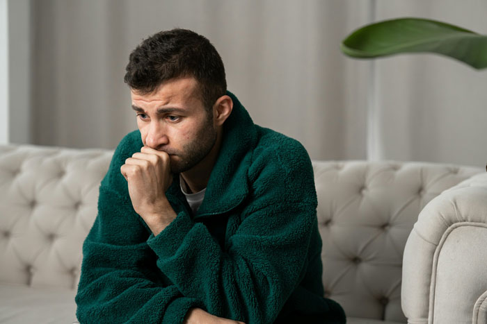 Man in green sweater looking worried on a couch, reflecting on moving in with girlfriend but refusing her apartment.