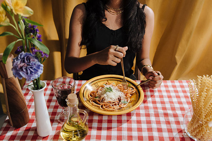 Woman eating spaghetti at a restaurant with a red and white checkered tablecloth and drinks nearby.