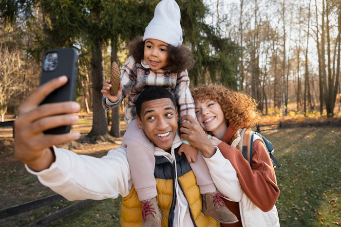 Happy family taking a selfie outdoors with child on father's shoulders during a fall day in the park.