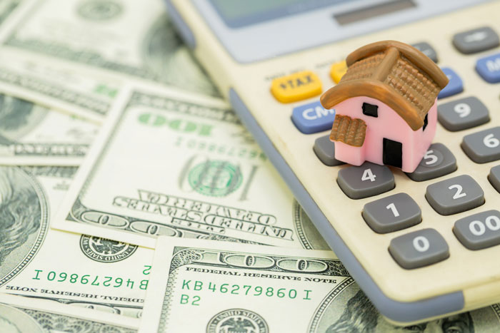 Miniature house on calculator buttons surrounded by dollar bills symbolizing rent and pay increase calculations.