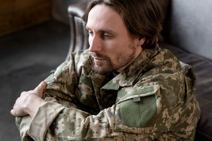 Man in military uniform sitting pensively on couch, reflecting on veteran bros pay increases and rent bill duty.