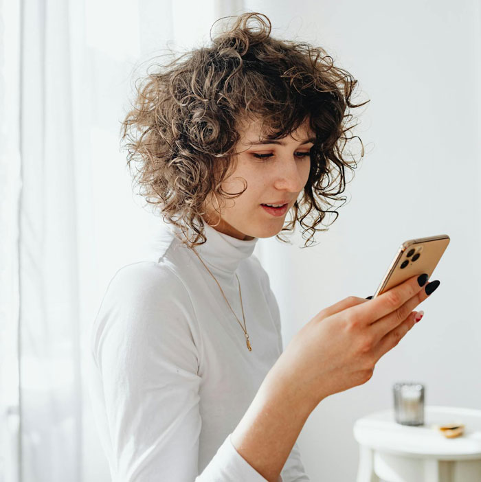 Woman with curly hair in a white turtleneck looking at her phone, reacting to drama at a groom&rsquo;s brother&rsquo;s wedding and baby shower.