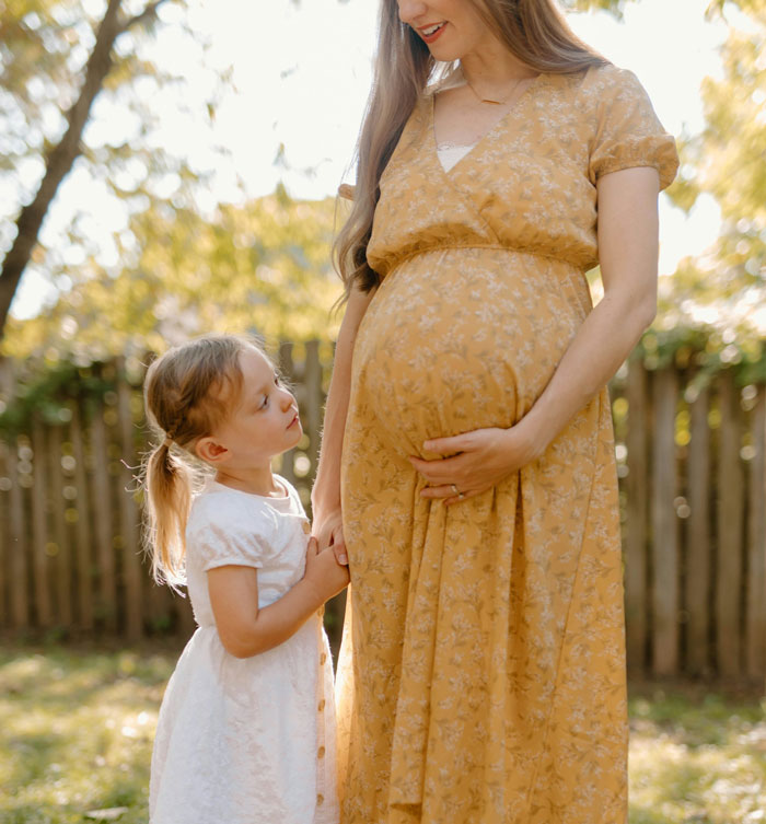 Pregnant woman in a yellow dress with a little girl holding her hand, representing baby shower and family moments outdoors.