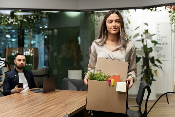 Office coordinator carrying a box of personal items while a pushy colleague watches in a modern office setting.