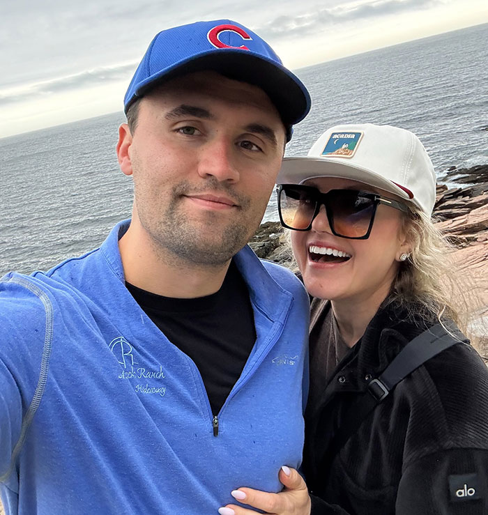 Two people posing outdoors by the ocean, with a woman wearing sunglasses and a cap, linked to Erika Kirk Epstein conspiracy.
