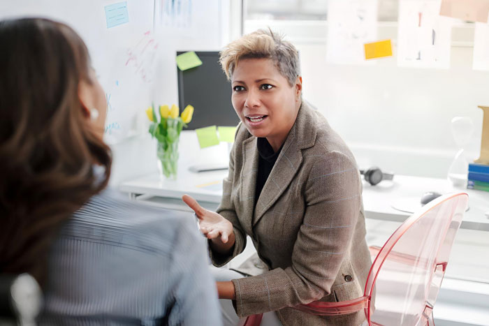 Woman in professional office attire talking seriously to colleague about frumpy clothes and workplace harassment.