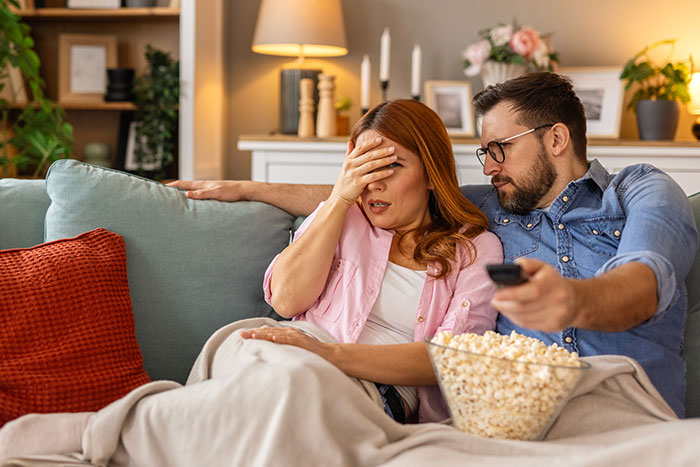Couple experiencing relationship drama on couch, woman upset while man holds remote, popcorn bowl nearby.