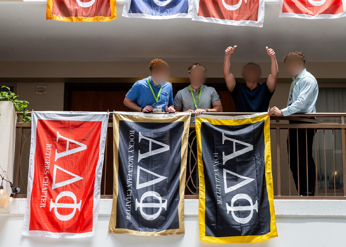 Four men stand behind fraternity flags hanging from a balcony during a fraternity hazing event interrupted by cops.