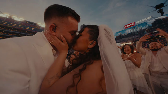 Couple sharing a kiss dressed in wedding attire during Bad Bunny’s halftime show at the Super Bowl in a large stadium.