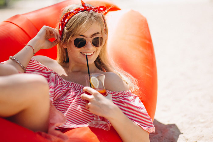 Young woman relaxing on orange inflatable at beach, enjoying a drink, representing cousin card fraud in vacation rental context
