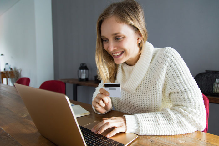 Young woman smiling and holding a card while using a laptop, illustrating cousin card fraud in vacation rental scams.