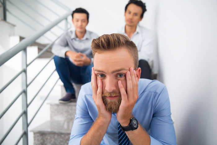Stressed man in blue shirt with coworkers behind him on stairs representing coworker reported HR gets fired situation Stressed man in blue shirt with coworkers behind him on stairs representing coworker reported HR gets fired situation