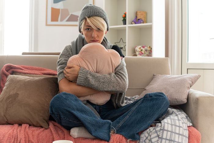 Young woman in a gray hat hugging a heart-shaped pillow on a couch, symbolizing secret concerns about sister responsibility.