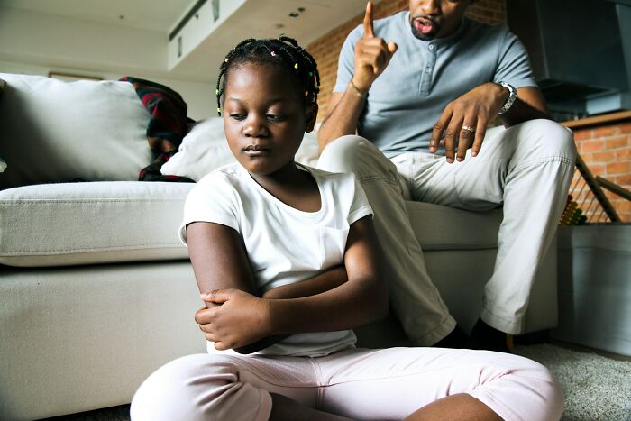 A young girl sitting with arms crossed looking upset while an adult man beside her gestures in a living room.