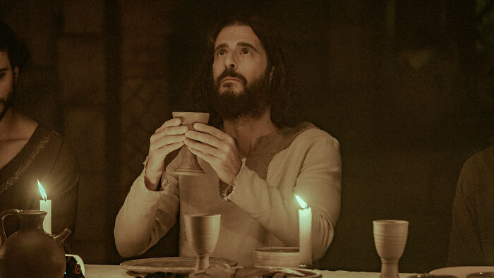 Scene from The Chosen with a man holding a cup at a dimly lit table, highlighting The Chosen Season 6 update by Dallas Jenkins.