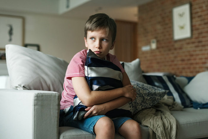 Boy sitting on couch hugging a pillow, looking upset inside a home where no kids are allowed.
