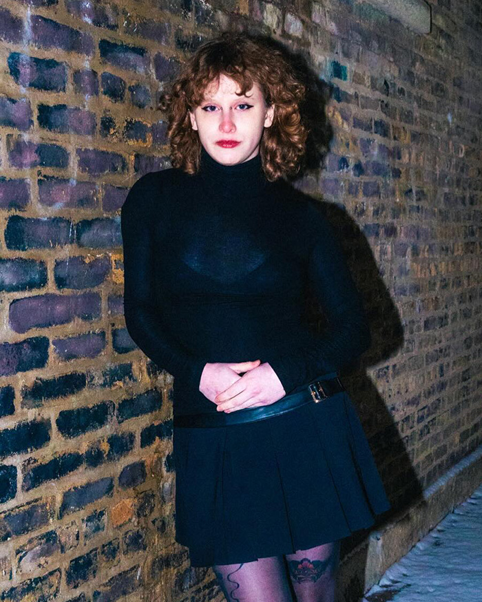 Indie musician standing against a brick wall at night, wearing a black turtleneck and skirt, highlighting concept copy accusations.