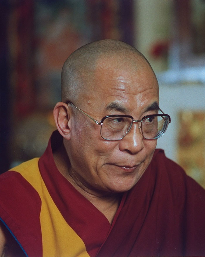 Dalai Lama wearing glasses and traditional robes, captured in a contemplative moment indoors with a blurred background.