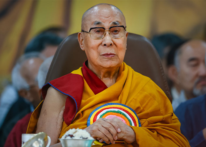 Dalai Lama seated in traditional robes with glasses, attending a formal event with people blurred in the background