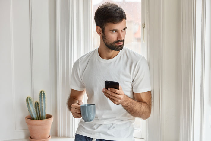 Nervous guy in white t-shirt holding coffee and phone, looking away thoughtfully by a window at home.