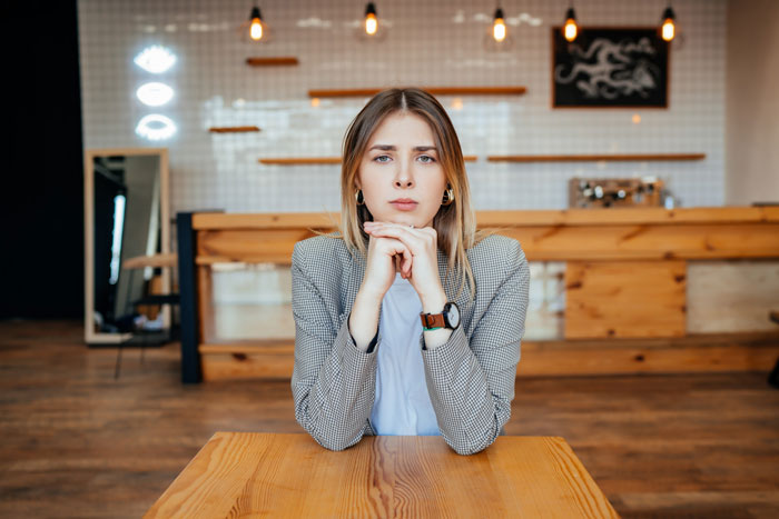 Young nervous woman sitting alone at a wooden table in a modern cafe, waiting for her first date with a friend chaperone nearby