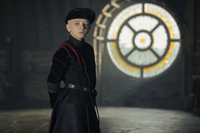 Young knight in dark medieval attire standing indoors near a stained glass window, related to A Knight Of The Seven Kingdoms season 2.