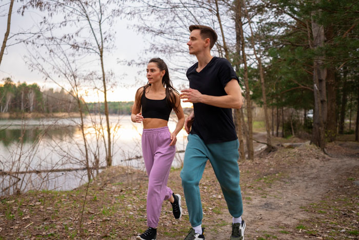 Couple running outdoors on a trail near a lake surrounded by trees, highlighting a husband&rsquo;s mandatory 5K run birthday activity.