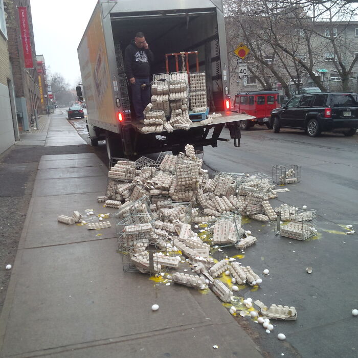 Man standing in truck bed next to spilled egg cartons on street, a funny photo showing an egg delivery fail.