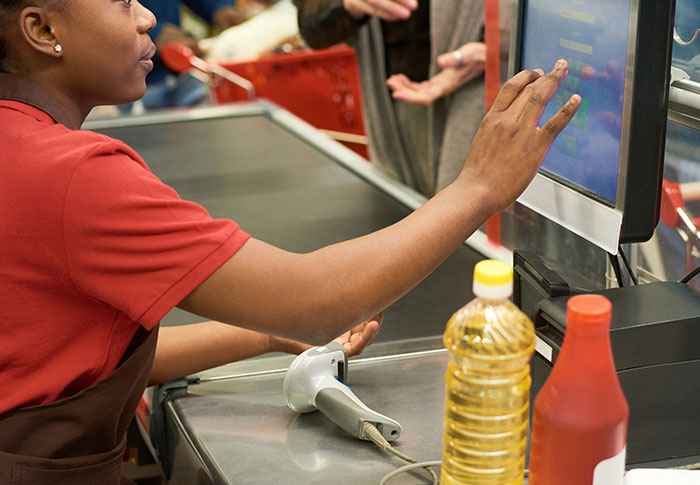 Grocery store cashier interacting with a touchscreen register while a customer gestures nearby, illustrating criminals shockingly bad at what they do.