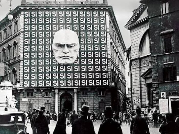 Black and white photo of a dystopian nightmare city scene with a large face sculpture on a building covered in repeated "SI" signs.