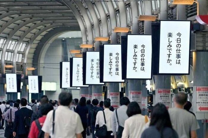 Crowded urban walkway with people passing under repetitive digital signs, evoking a dystopian nightmare scene.